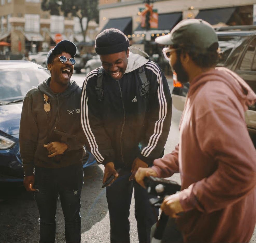 Three friends laughing together on a sunny street, one holding a scooter and others wearing casual hoodies and jackets.