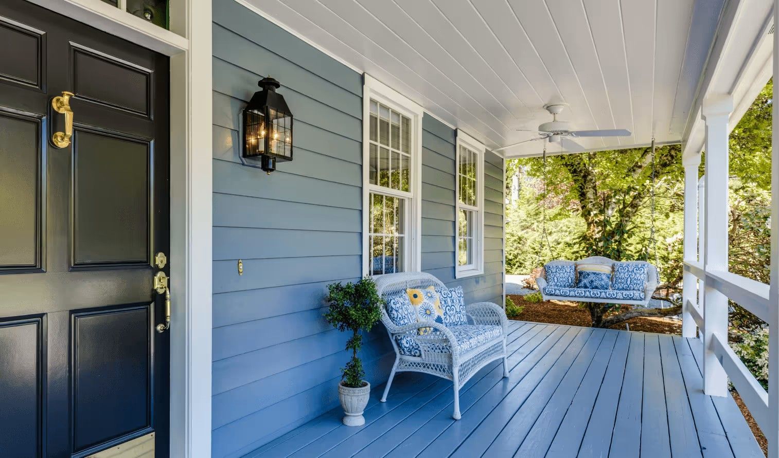 A blue-painted front porch with a dark blue door, white wicker furniture, a ceiling fan, and a hanging porch swing.