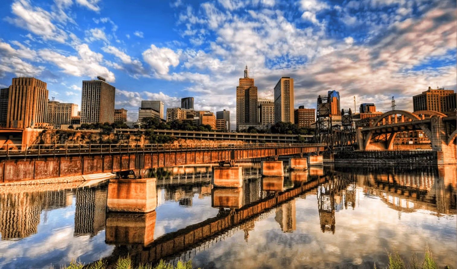 A skyline of St. Paul, Minnesota, at sunset with a railroad bridge and its reflection in the Mississippi River.
