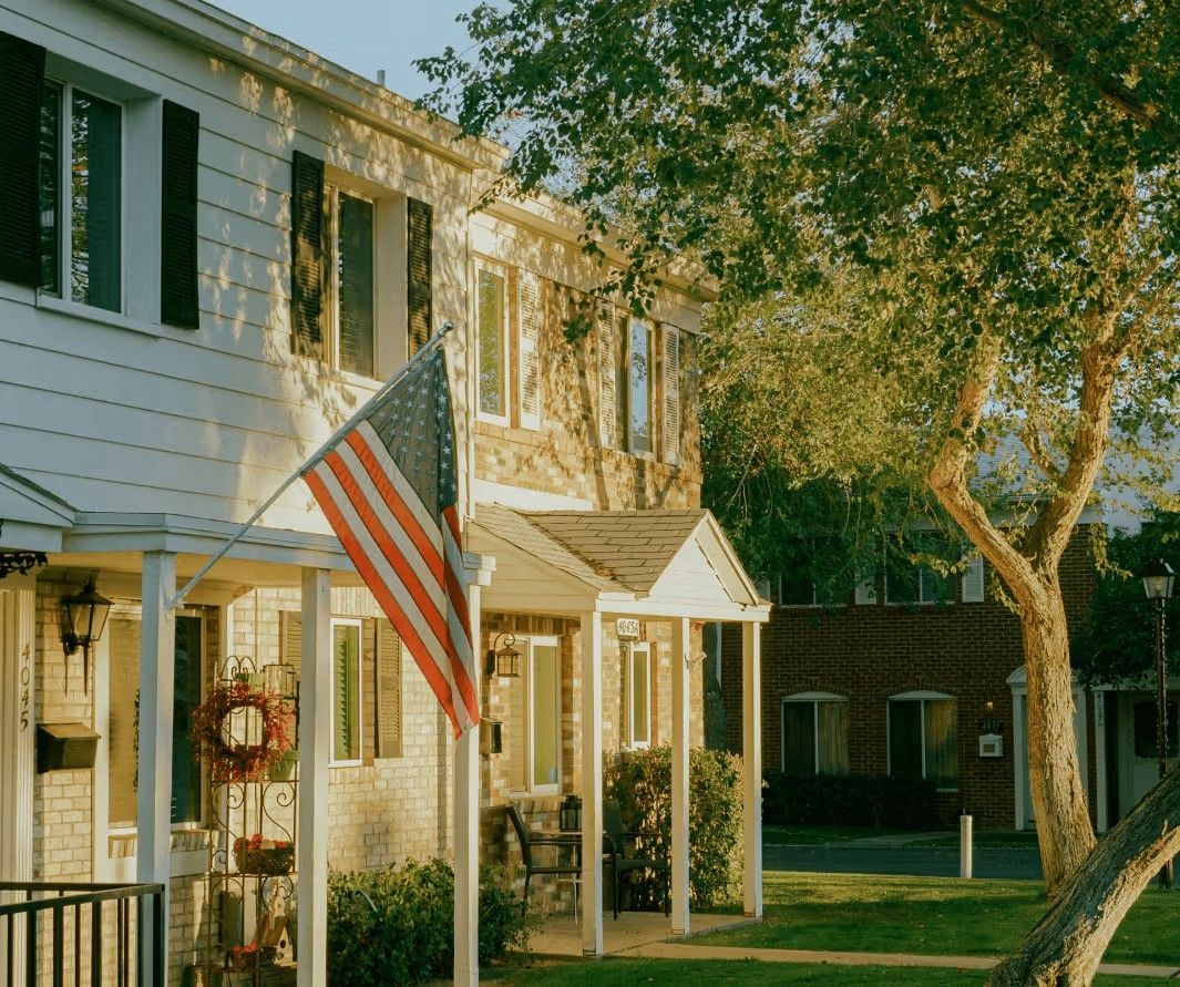 An American flag hangs from the porch of a white two-story suburban townhouse on a sunny day.
