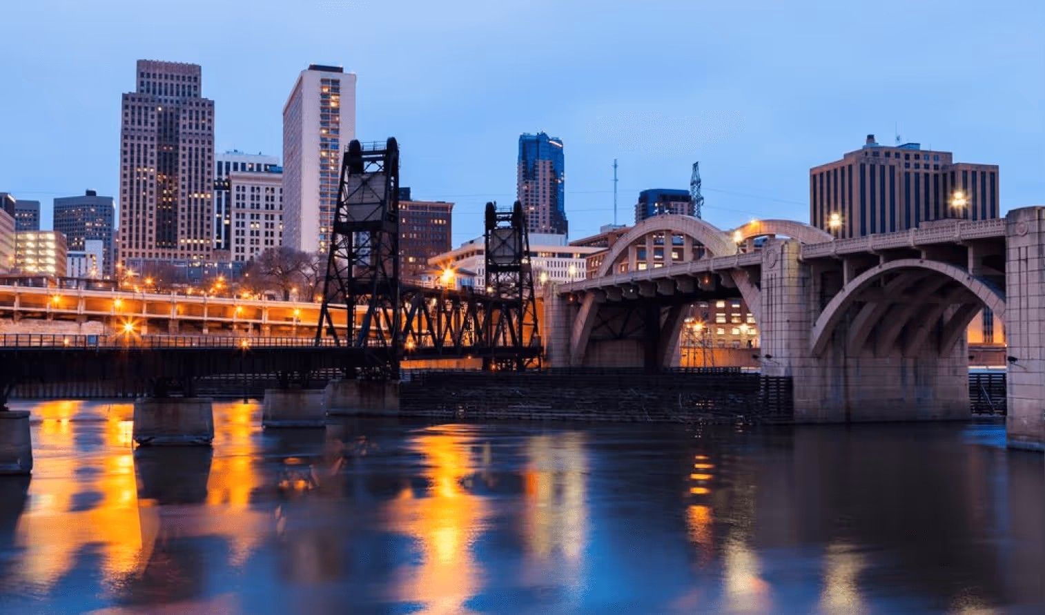 City skyline at twilight, featuring an arched stone bridge and a steel vertical-lift bridge spanning a river.