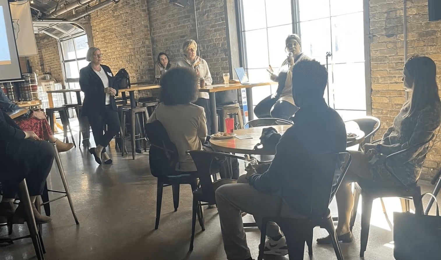 A group of people sit at tables in a rustic, brick-walled room during an indoor professional presentation or workshop.