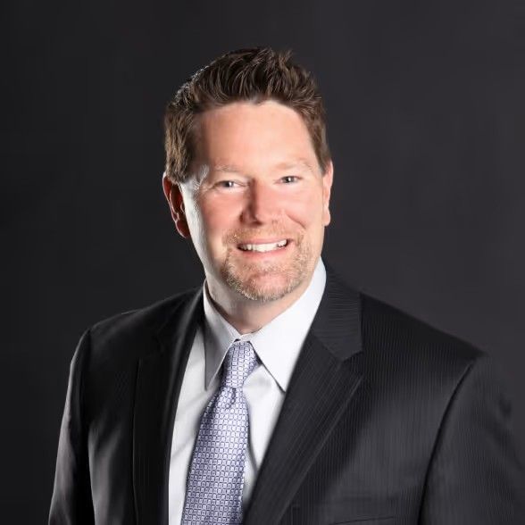 Professional headshot of a smiling person wearing a dark suit, white collared shirt, and patterned tie against a black background.