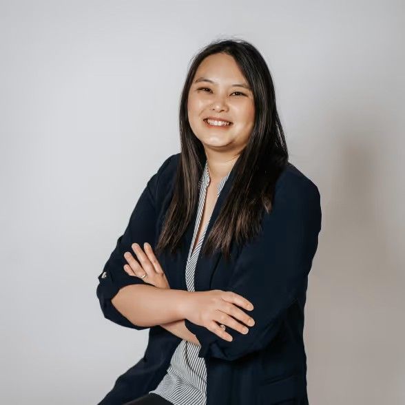 A smiling person with long dark hair, wearing a navy blazer over a patterned top, sits with their arms crossed.