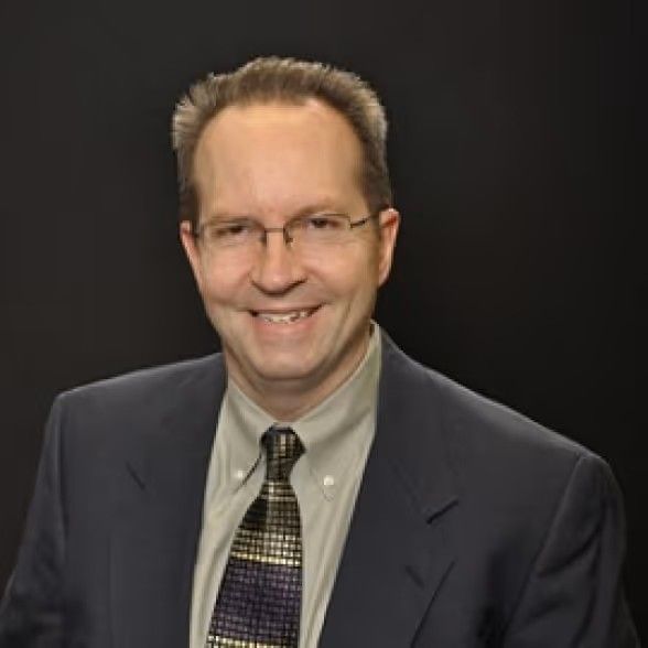 A smiling man in a suit, light-colored shirt, and patterned tie against a plain black background.