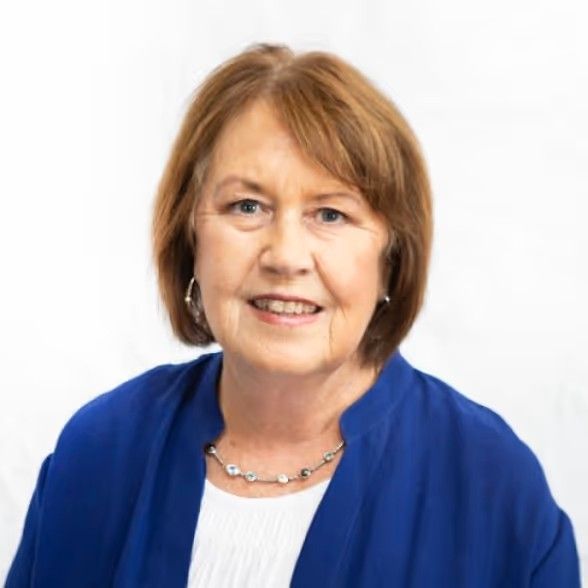 A smiling person with auburn hair wearing a blue jacket and a silver necklace against a white background.