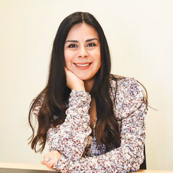 A smiling person with long dark hair wears a floral print top, resting their chin on their hand at a desk.