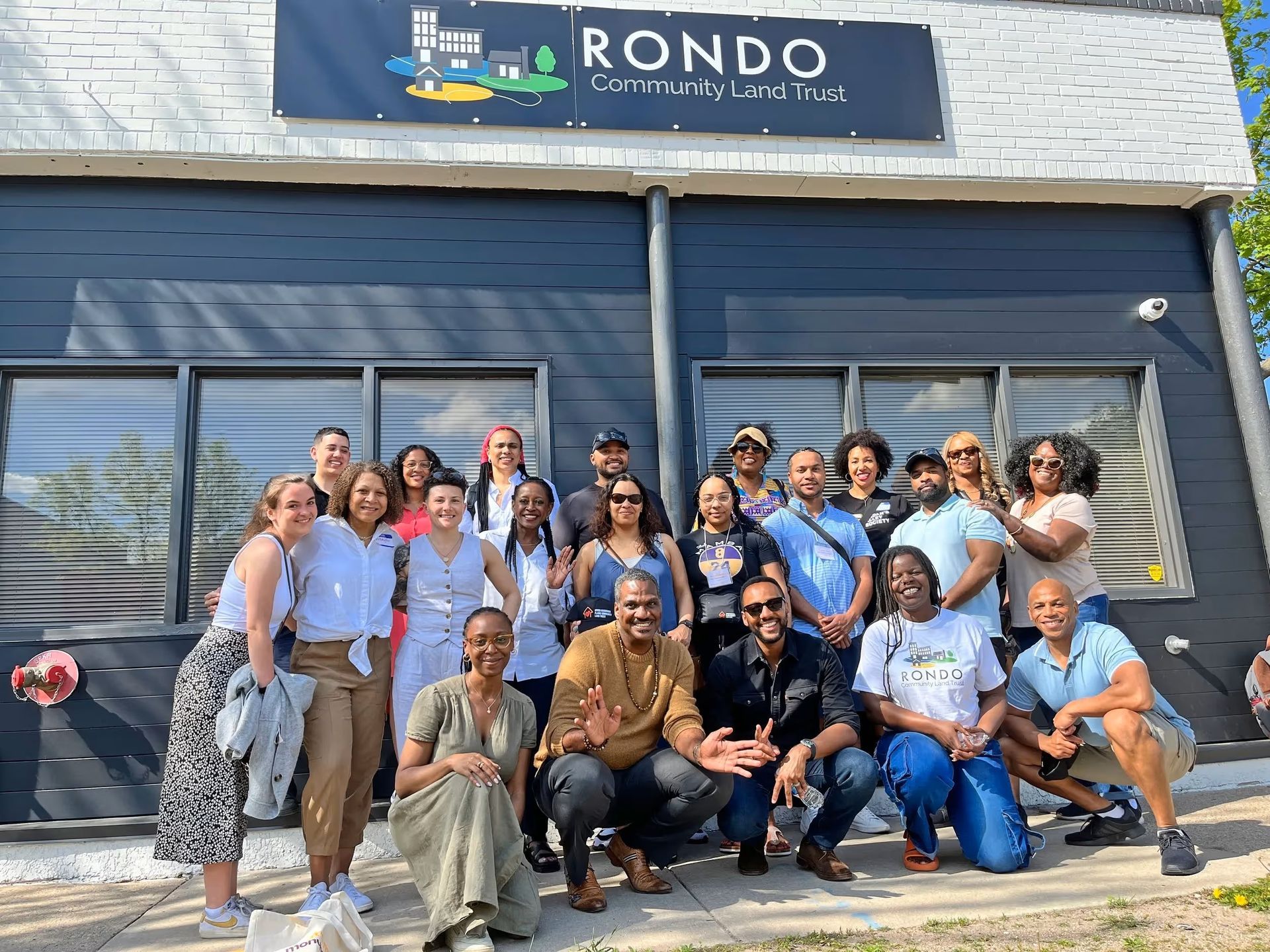 A group of people standing and smiling in front of the Rondo Community Land Trust building on a sunny day.