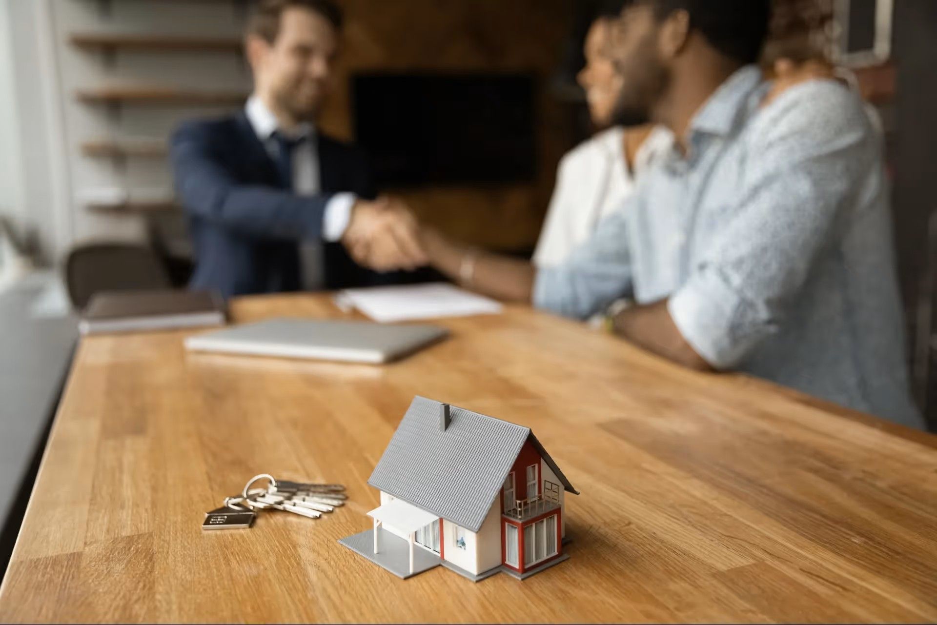 A miniature house and keys sit on a desk in the foreground as people shake hands in the blurred background.