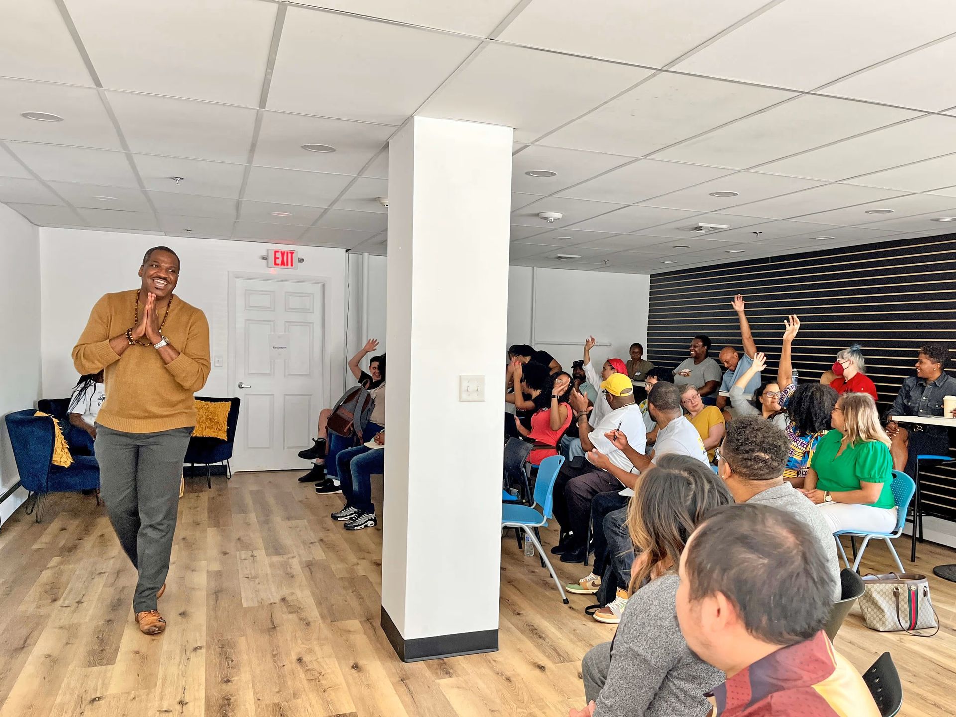A presenter stands before an audience in a bright, modern room while several people raise their hands to participate.