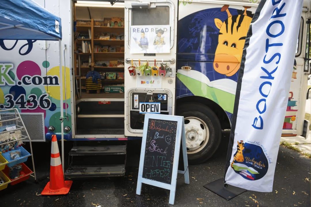 A mobile bookstore van with a giraffe mascot, an open door revealing shelves of books, an open sign, and a chalkboard sign.