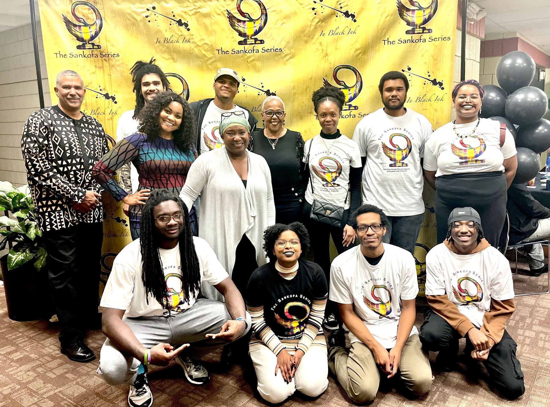 A group of 15 people pose for a photo in front of a yellow banner featuring a circular logo.