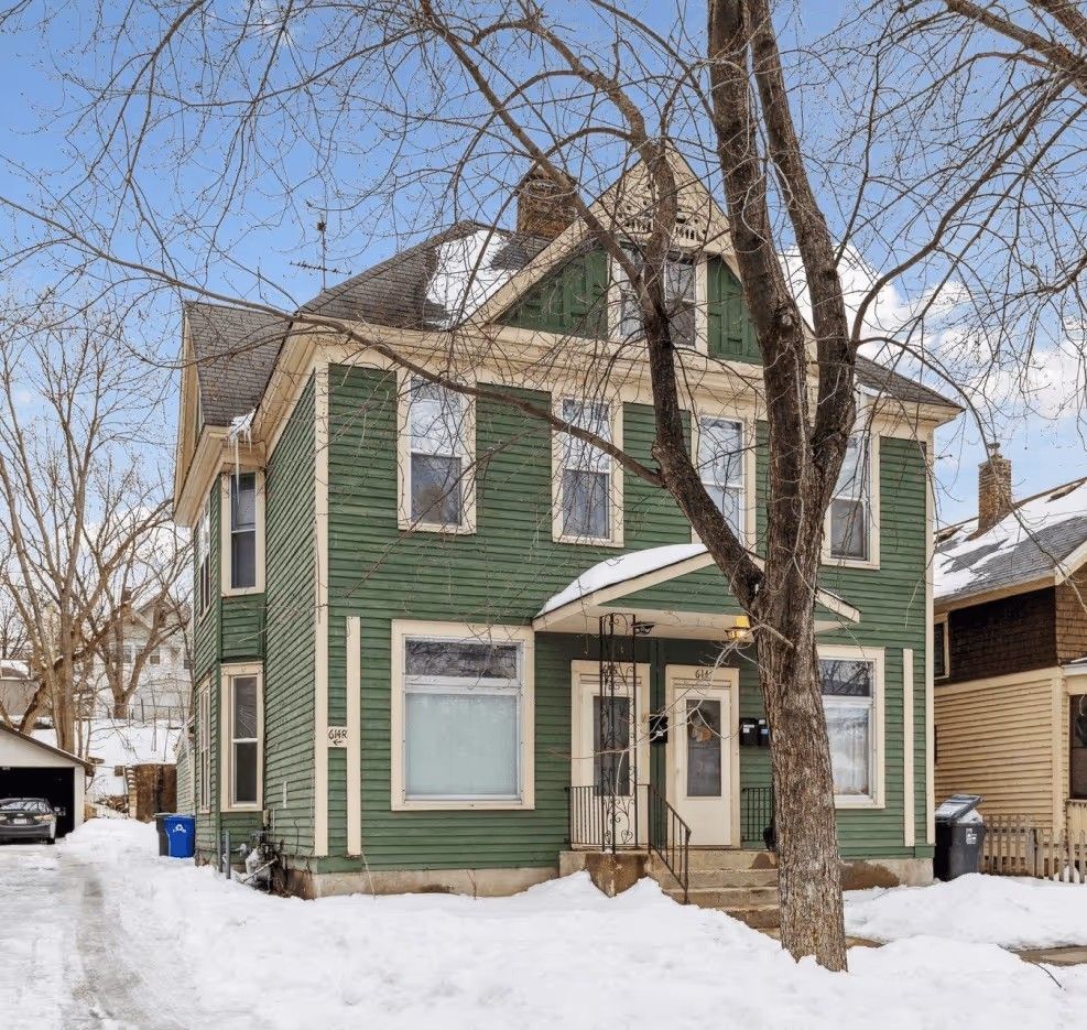 A two-story, green-sided Victorian-style house with cream trim and snow-covered ground on a bright, winter day.