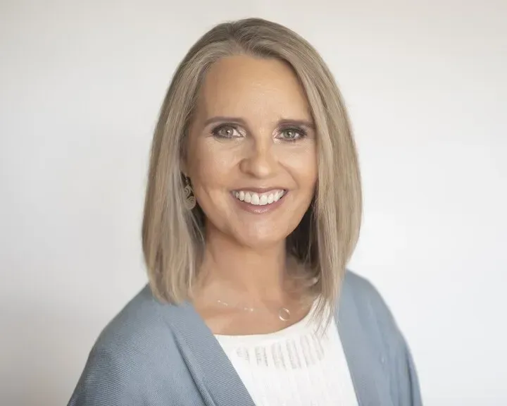 A person with shoulder-length blonde hair, wearing a white top and blue cardigan, smiling against a white background.