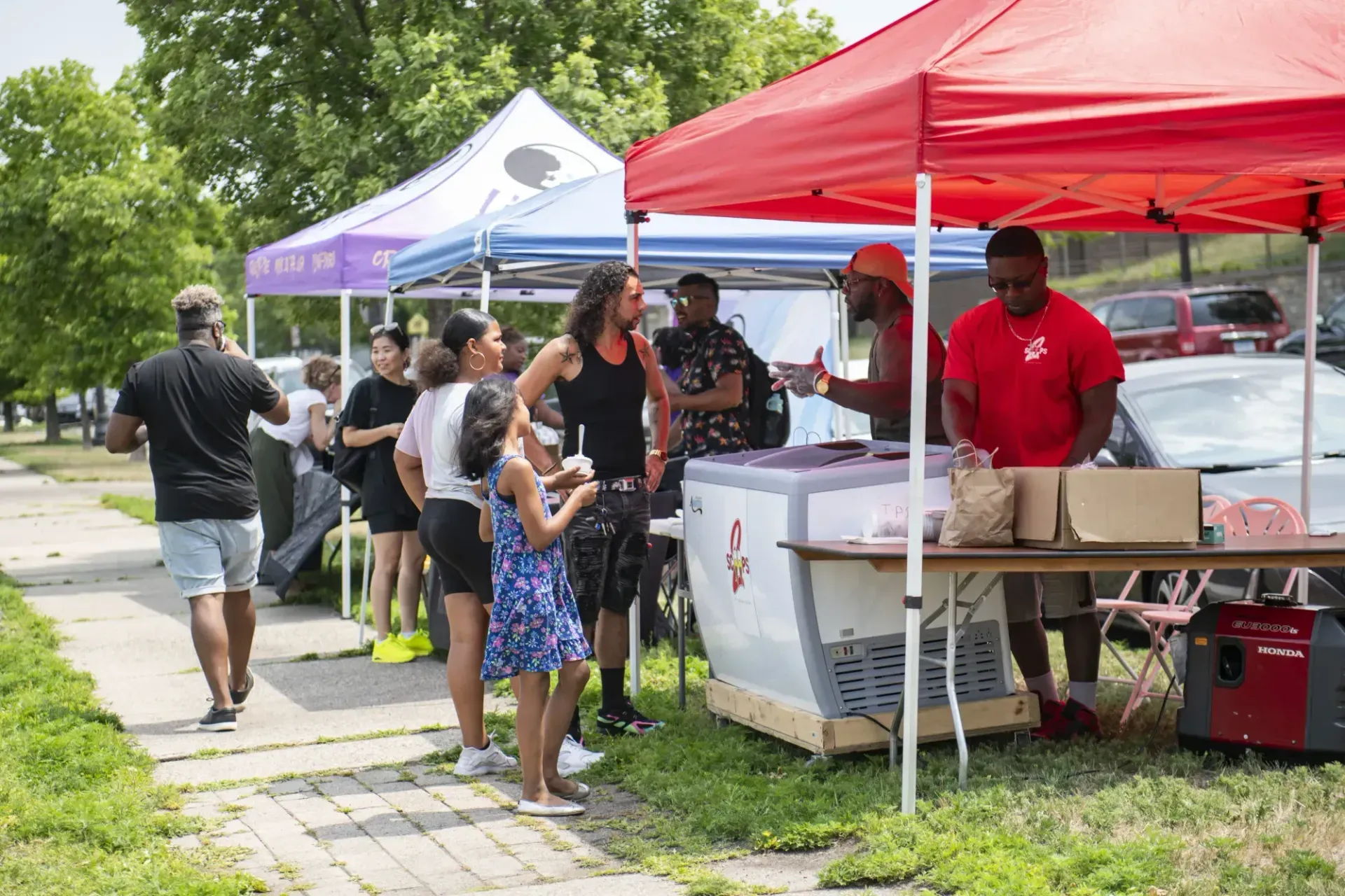 People stand and chat under red and blue pop-up tents at an outdoor community event with a cooler and table setup.