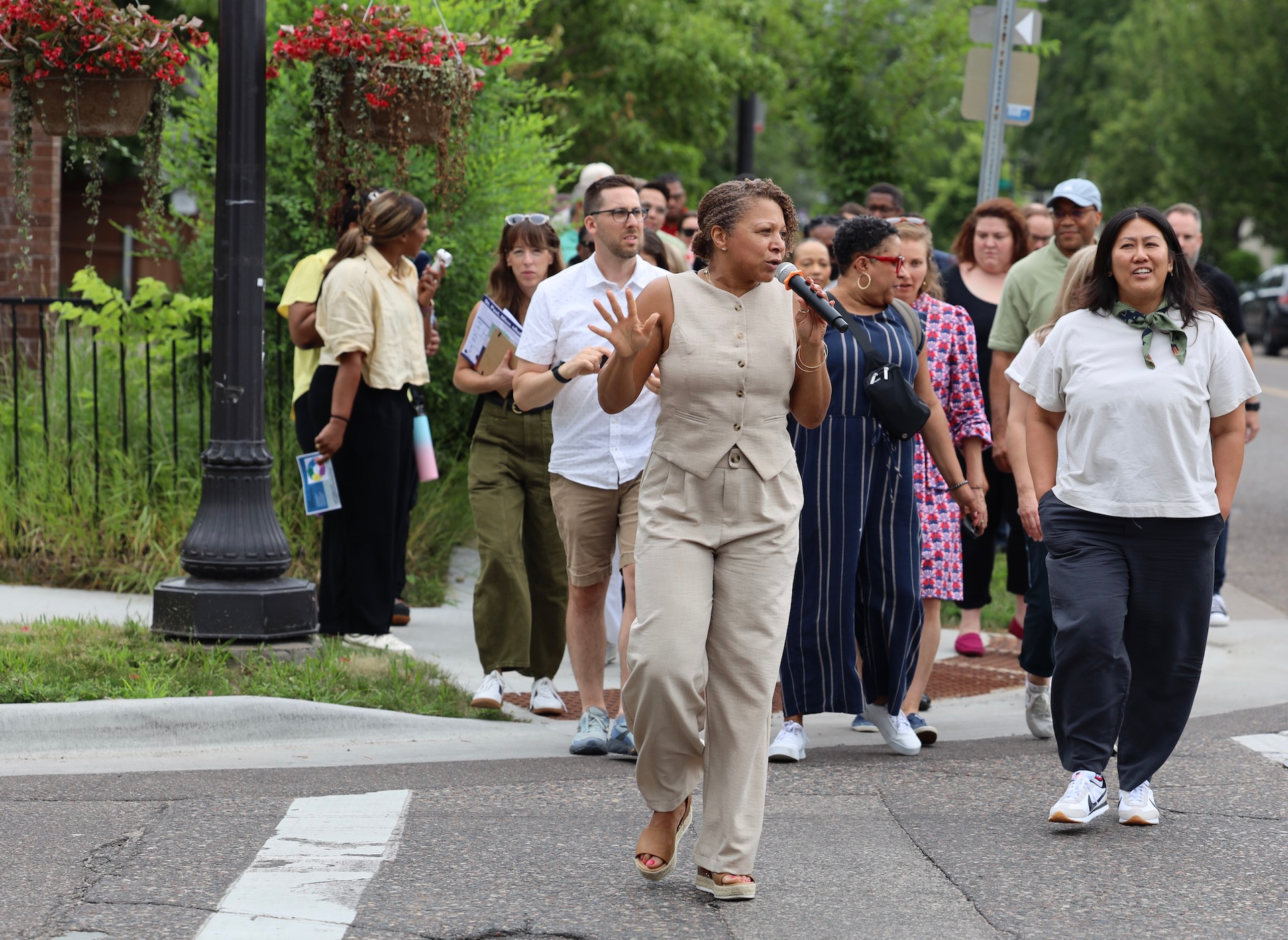 A group of people walks across a city street; one person in the center speaks into a microphone while gesturing.