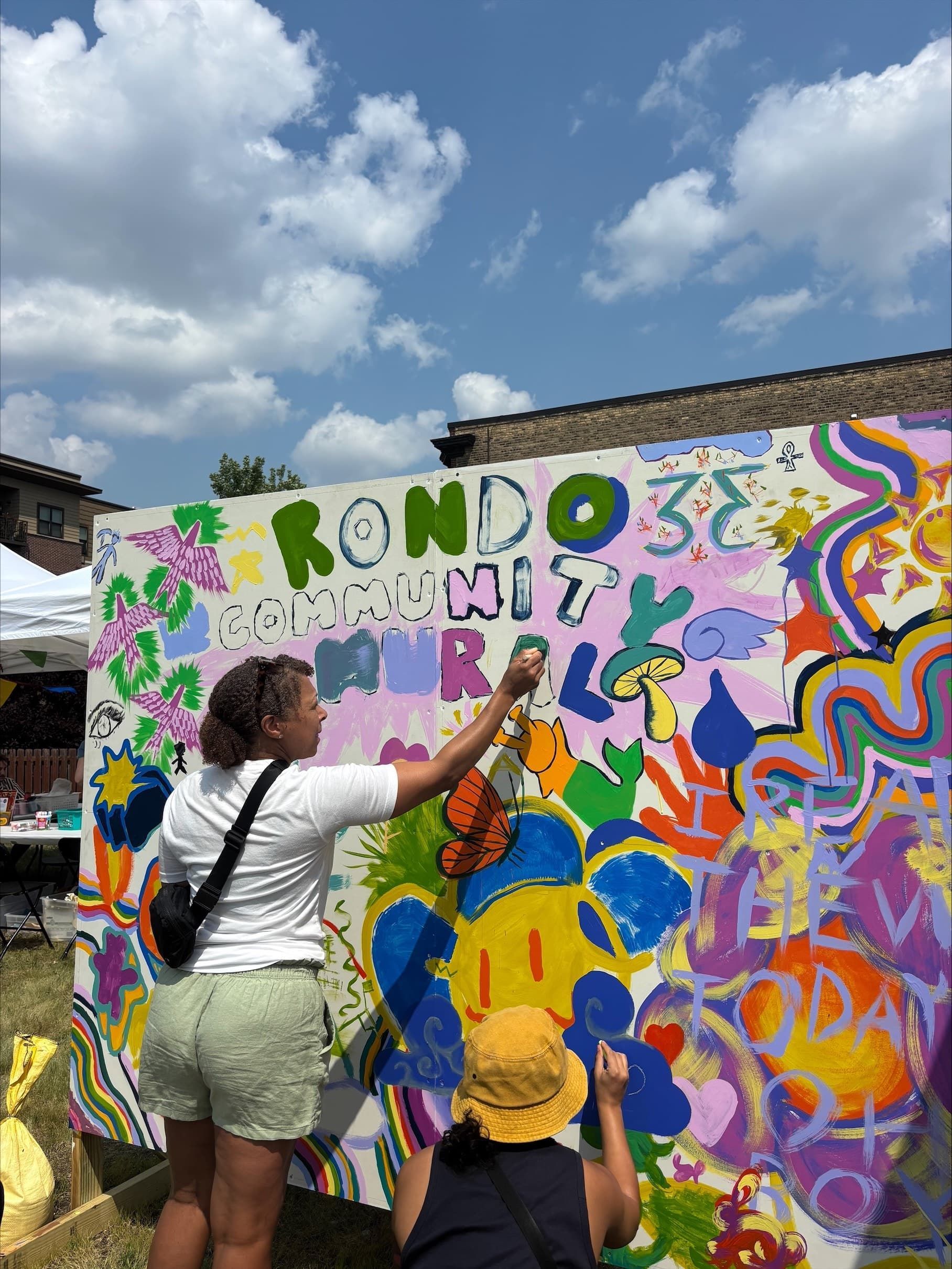 Two people painting a colorful community mural outdoors on a sunny day.