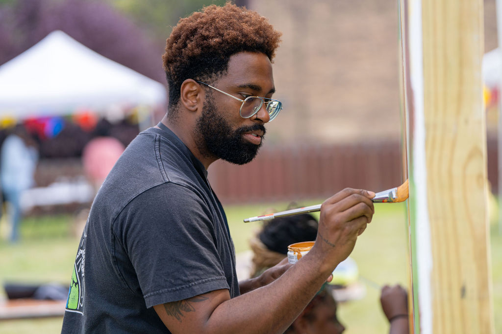 A person with a beard and glasses focuses intently while painting on a wooden board outdoors.