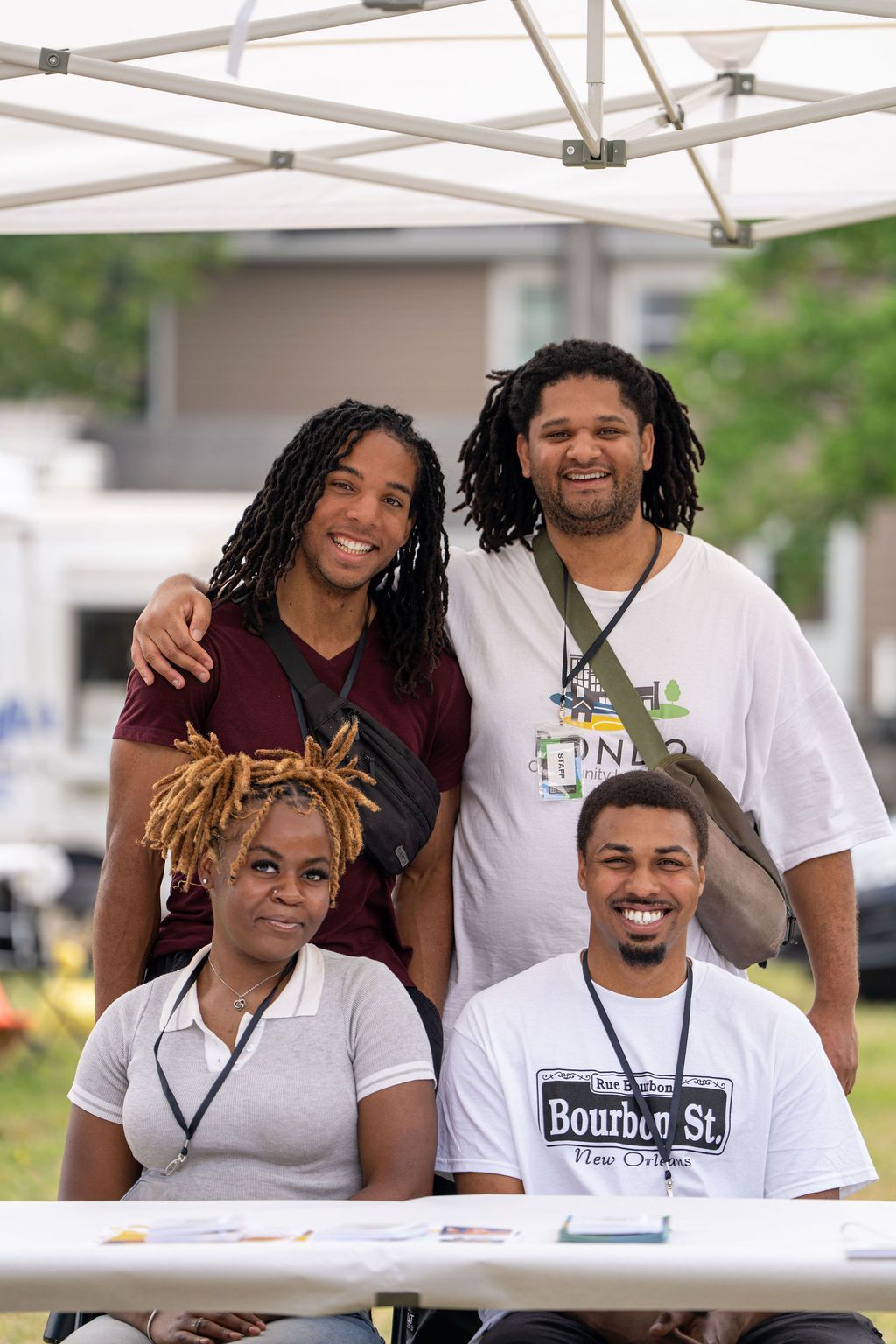 Four people pose under a white tent canopy at an outdoor event, standing and sitting behind a white table.