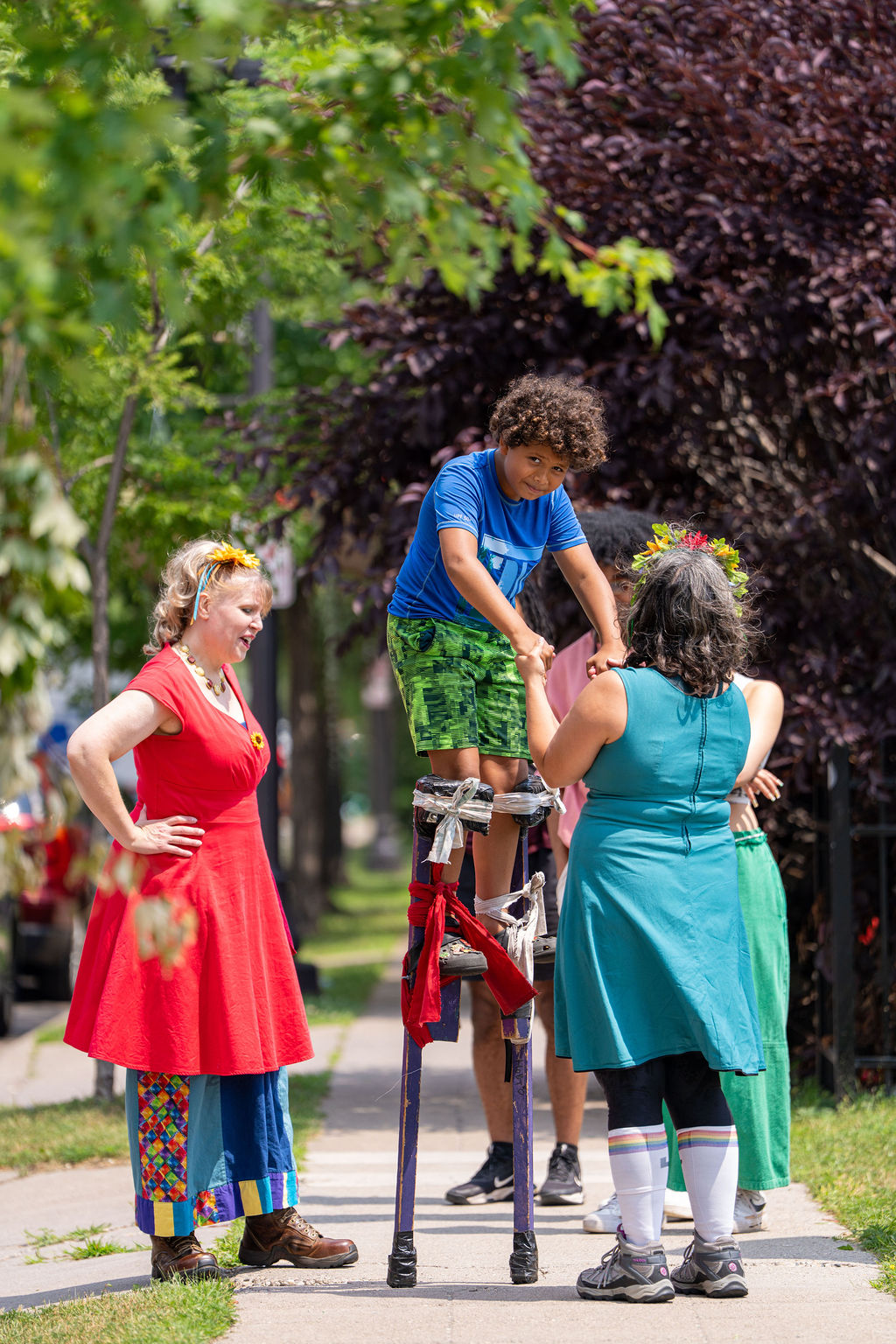 A child on stilts holds hands with a person in a teal dress, while another person in a red dress watches on a sidewalk.