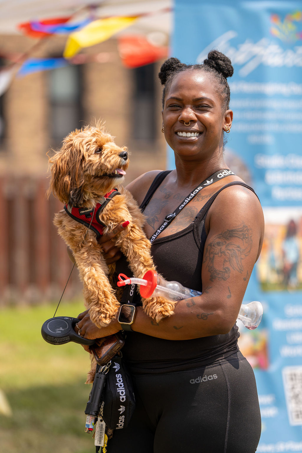 A smiling person holds a small, fluffy tan dog in a harness outdoors near a blue informational banner.