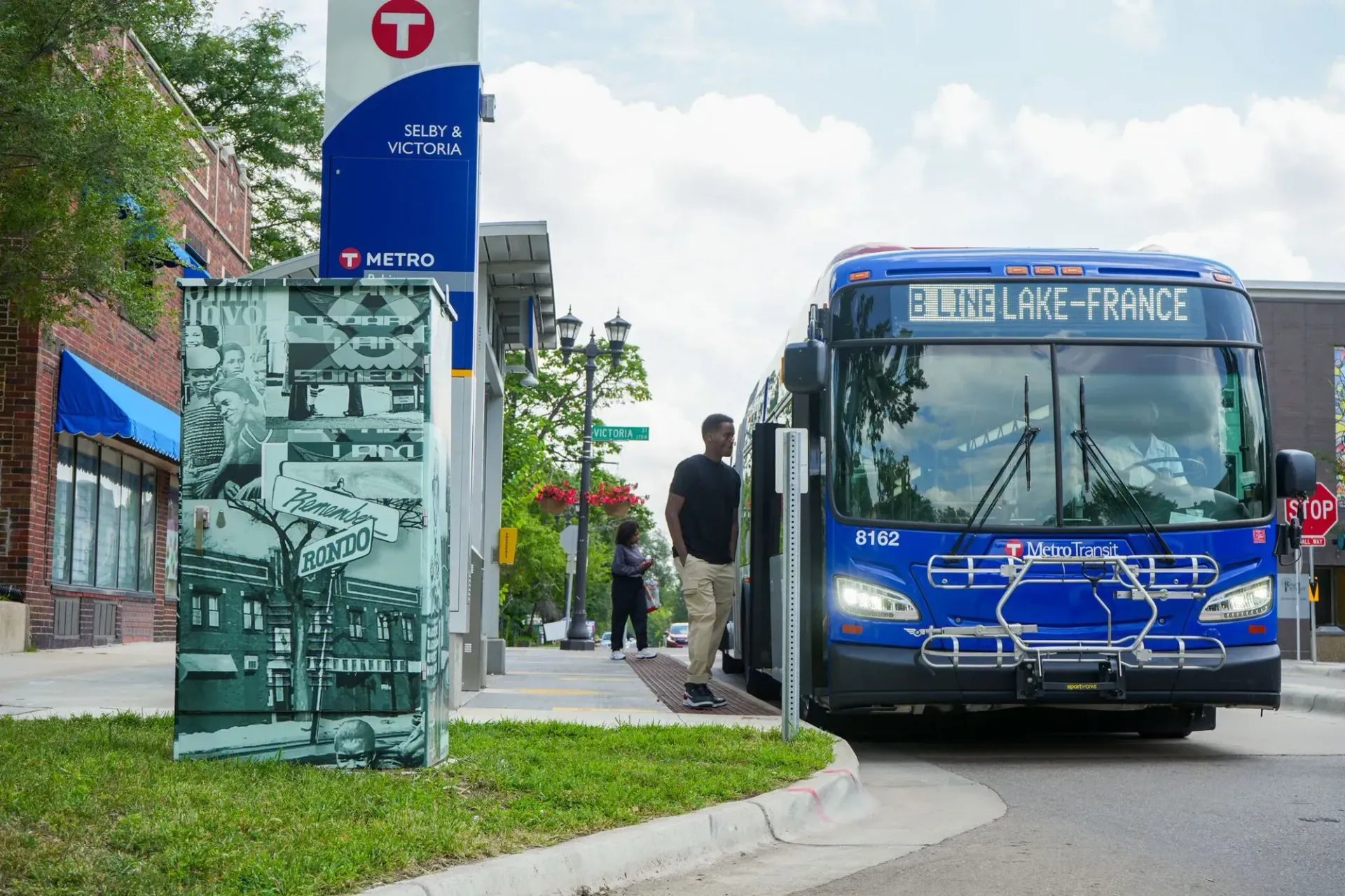 A blue city bus is stopped at a Metro Transit bus stop next to a decorated utility box on a sidewalk.