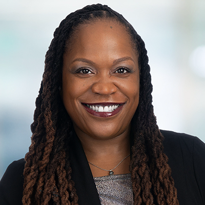 Headshot of a smiling person with dark, long, braided hair, wearing a black blazer over a metallic top and a necklace.