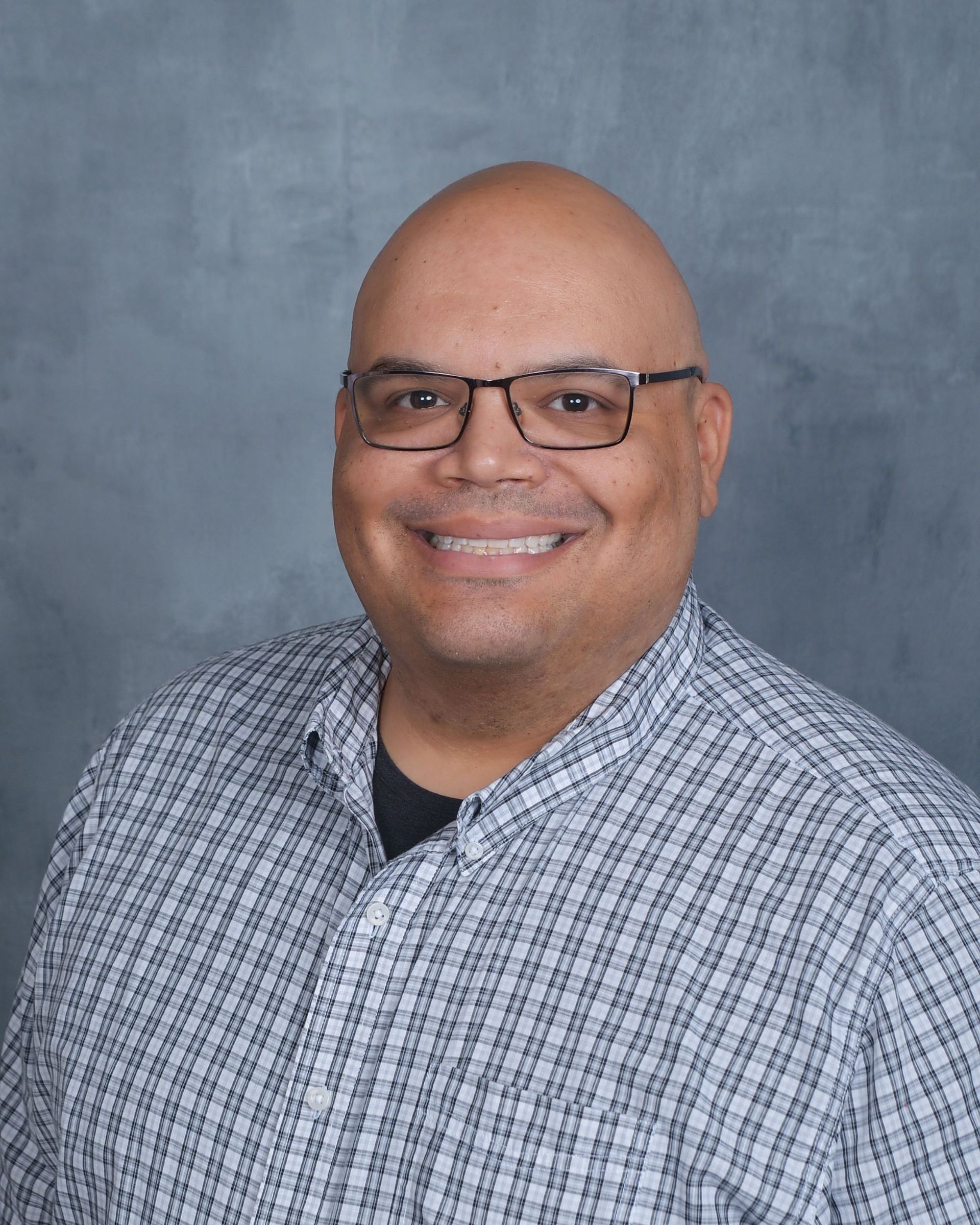 A person wearing glasses and a plaid button-down shirt smiles in front of a mottled gray background.