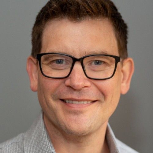 A smiling person with brown hair and dark-rimmed glasses wearing a light gray patterned shirt, against a gray background.