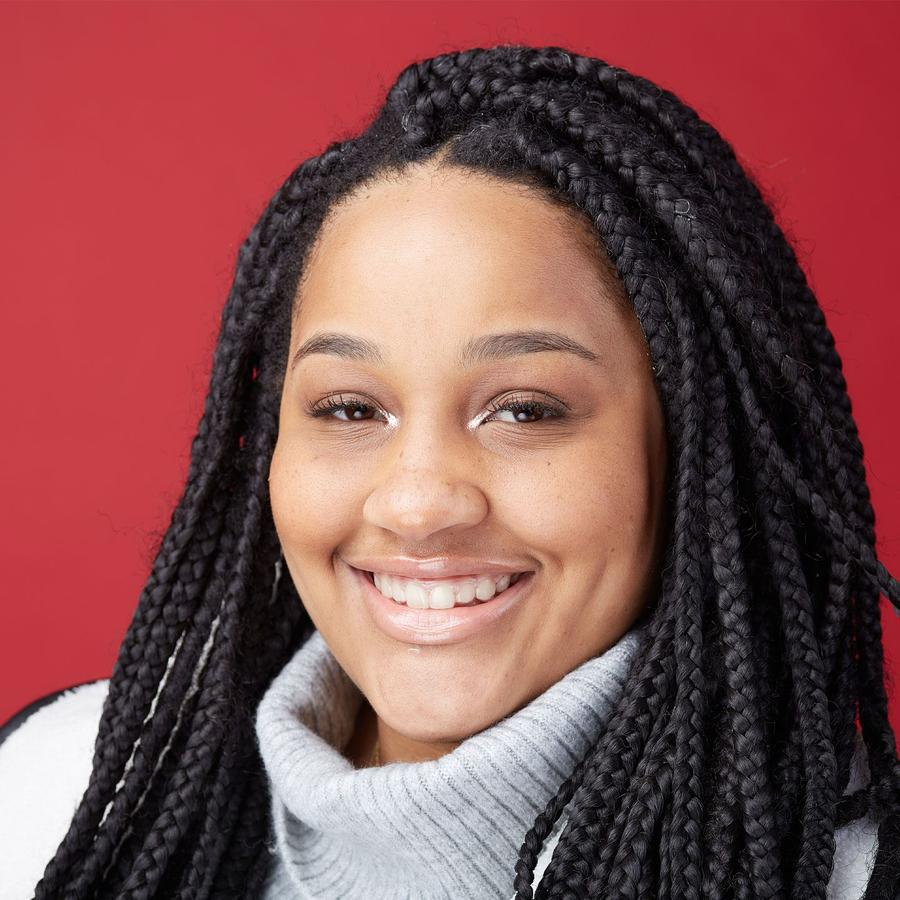 A person with braided hair smiling, wearing a grey turtleneck sweater against a solid red background.