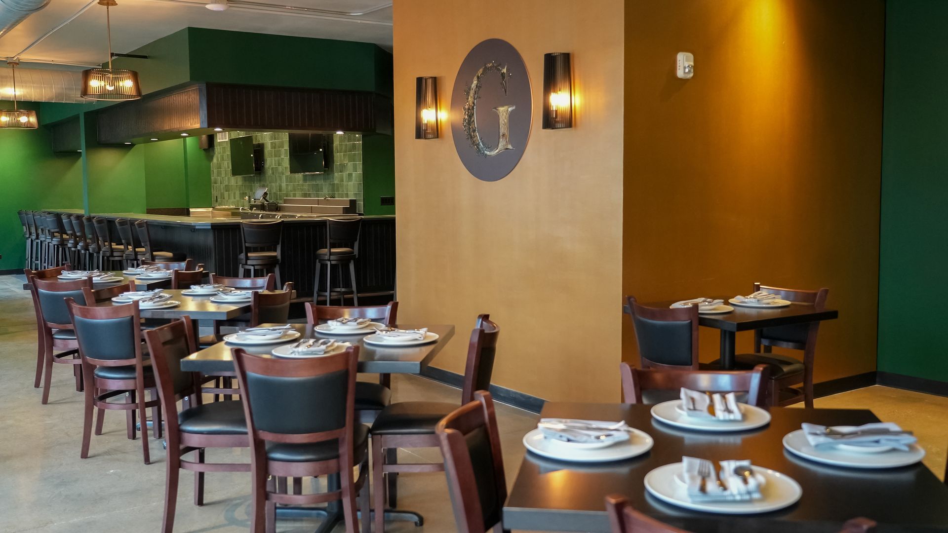 An interior view of a restaurant with green walls, dark wooden tables and chairs, and a bar area in the background.