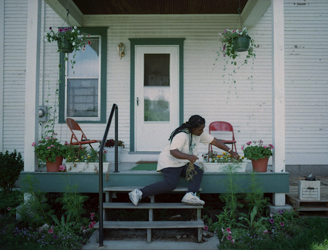 A person sits on porch steps, tending to a long planter box filled with flowers in front of a white house.