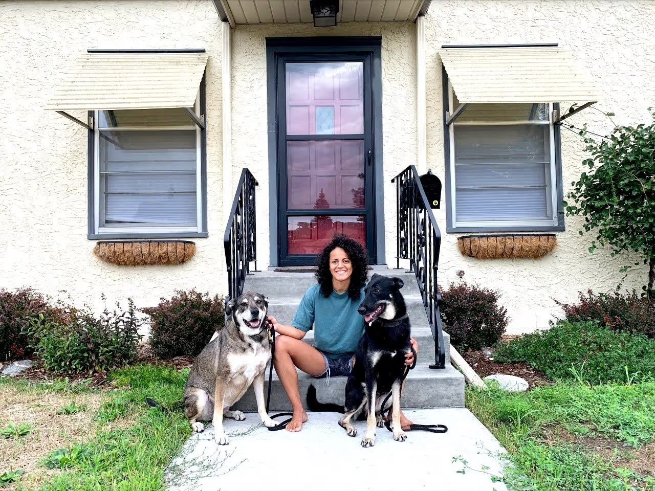 A person sits on stone steps in front of a house, flanked by two large dogs, with a smiling expression.