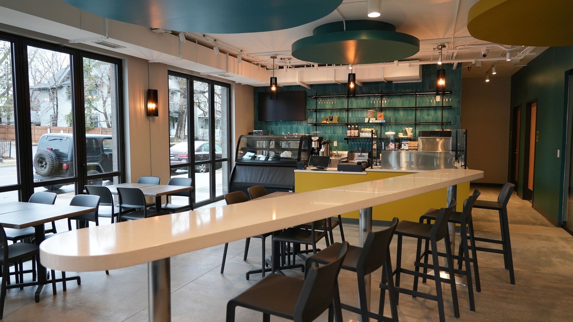 Modern, brightly lit cafe interior featuring a long, white quartz bar, yellow counter, and stools against glass windows.
