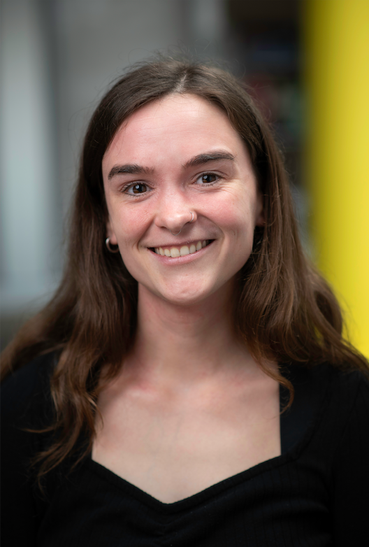 A smiling person with long brown hair wearing a black sweater, posed in front of a white shelving unit with decor.