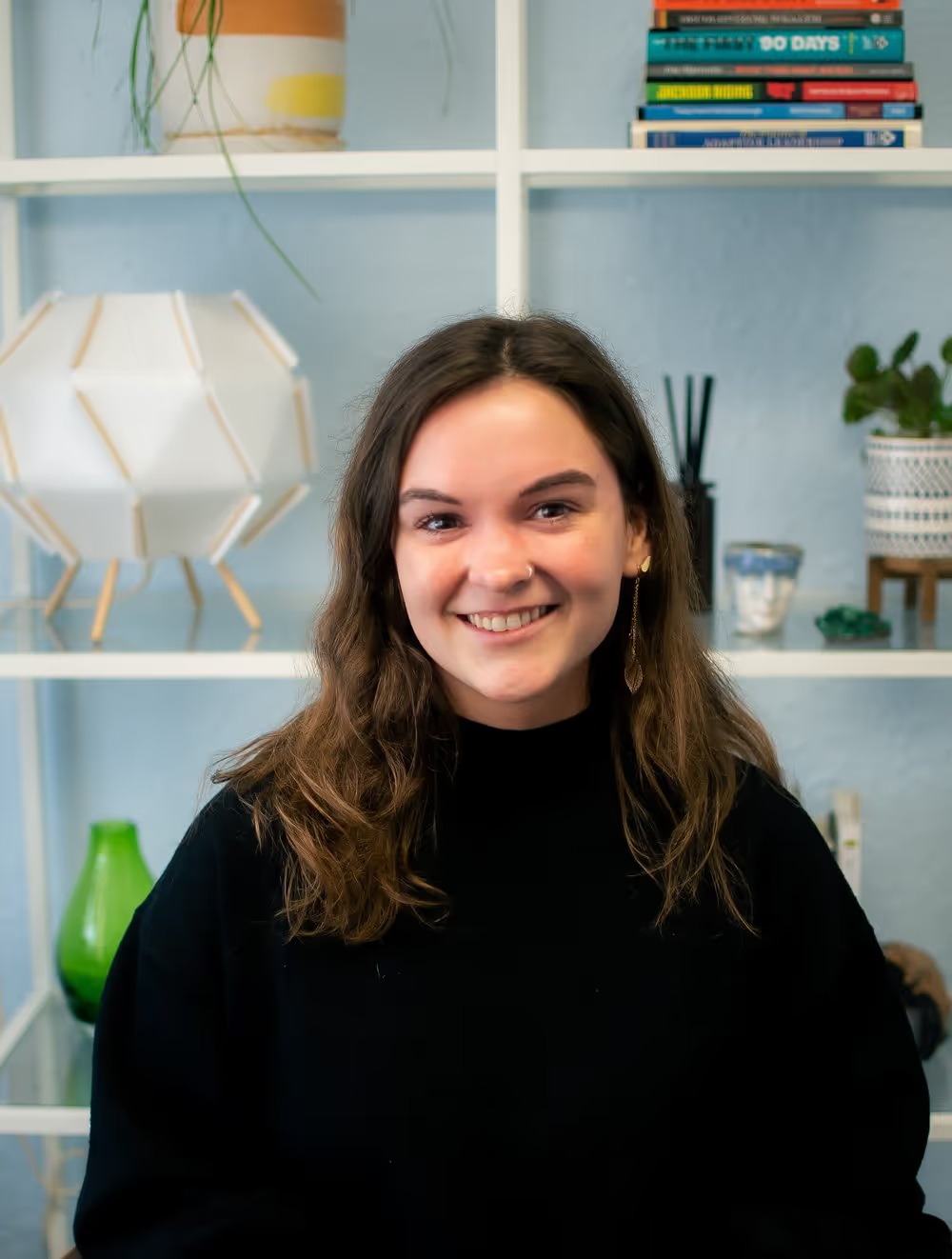 A smiling person with long brown hair wearing a black sweater, posed in front of a white shelving unit with decor.