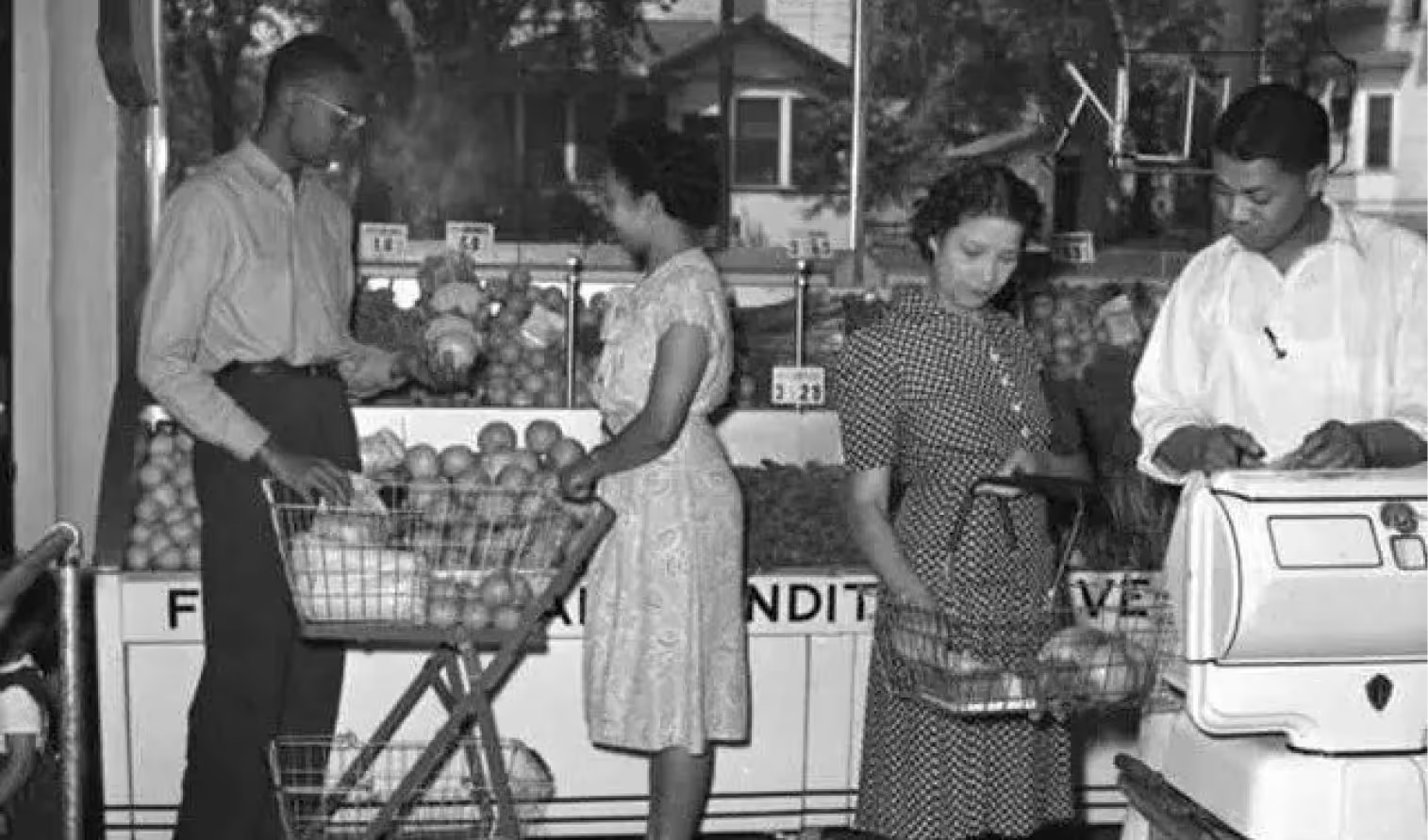 Customers shopping for produce in a grocery store as an attendant prepares to weigh items on a scale.