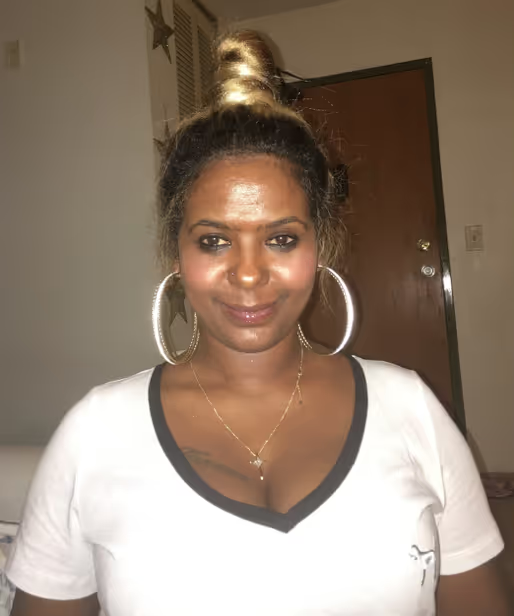A person with a blonde topknot, large hoop earrings, and a white V-neck shirt, posing indoors.