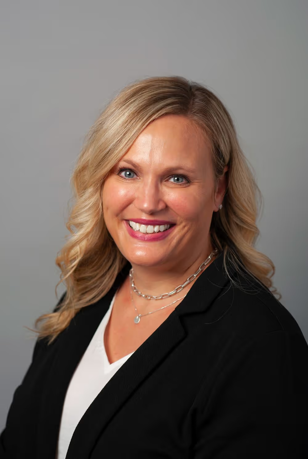 Headshot of a smiling person with blonde hair, wearing a black blazer over a white shirt, against a gray background.