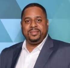 A man with a goatee wearing a white shirt and dark blue suit jacket against a blue geometric background.