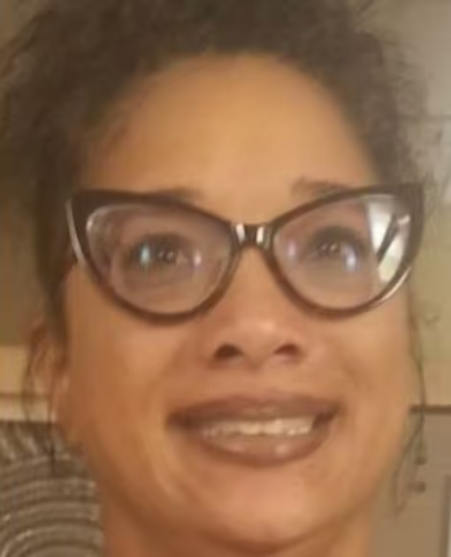 A close-up portrait of a smiling person with curly dark hair wearing dark cat-eye glasses.