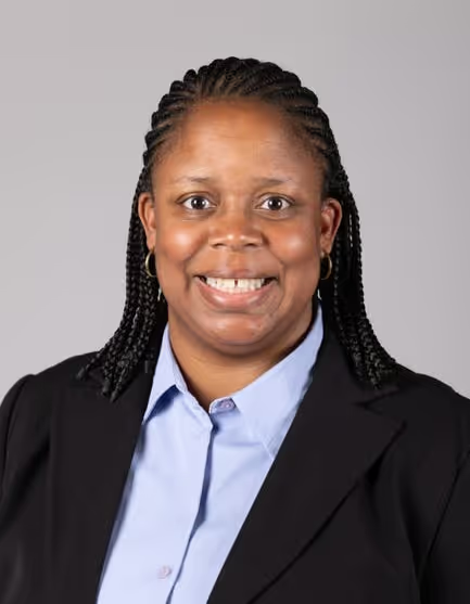 A person with braided hair, wearing a light blue collared shirt and black blazer, smiles against a plain gray background.