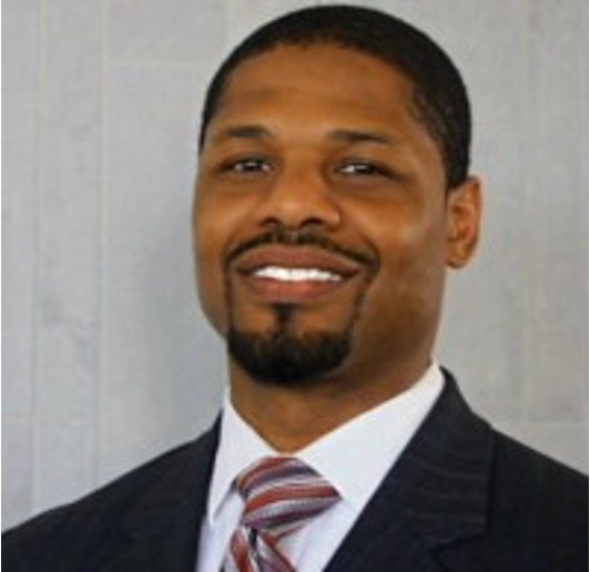 A person smiling, wearing a dark suit, white collared shirt, and a striped tie, against a light gray background.