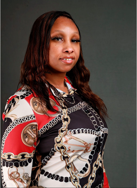 A person with long brown hair poses against a gray background wearing a top with a bold red, black, and gold chain print.