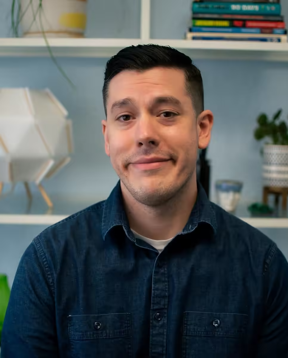 A person in a denim shirt smiles slightly against a light blue wall with shelves displaying books, plants, and decor.