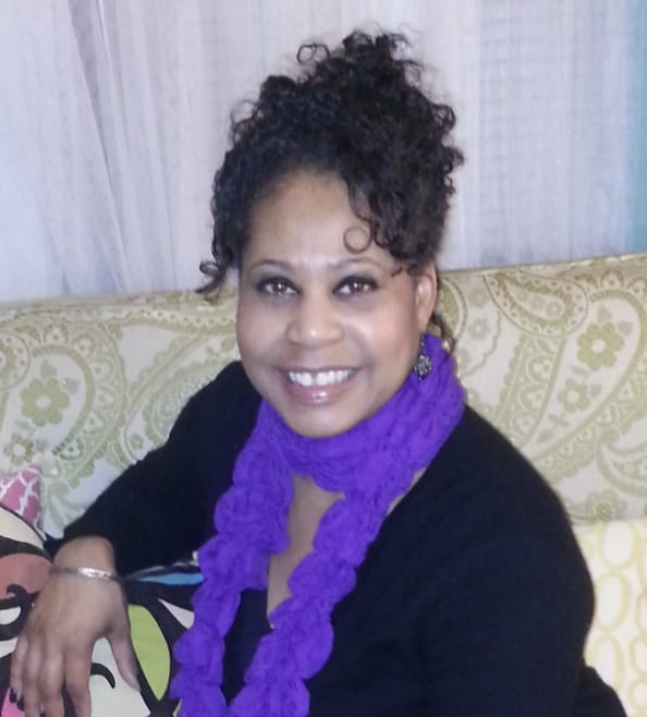 A smiling person with curly dark hair wearing a black top and a bright purple ruffled scarf, sitting on a patterned sofa.