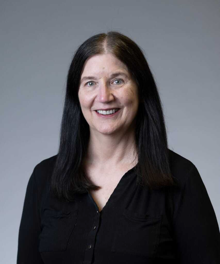 A person with shoulder-length dark hair smiling while wearing a black button-up shirt against a grey background.