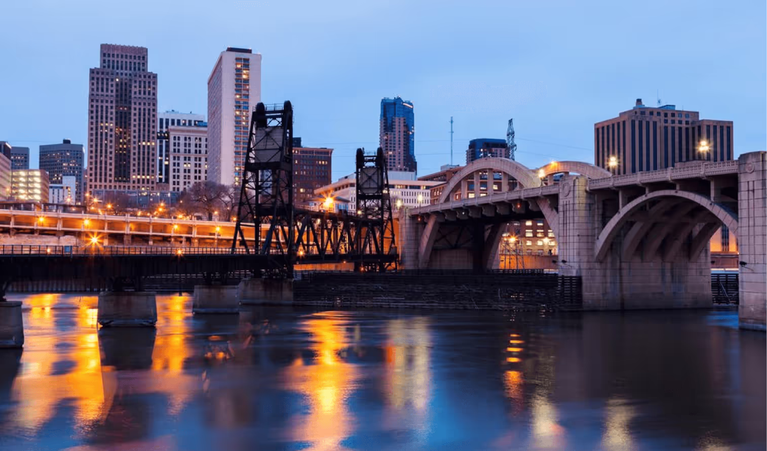 The Minneapolis skyline at twilight, featuring the historic Stone Arch Bridge and Hennepin Avenue Bridge over the river.