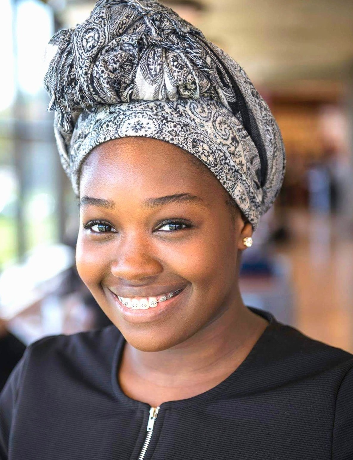 A smiling person with a patterned headwrap and black zip-front top, captured in a softly lit, professional portrait.