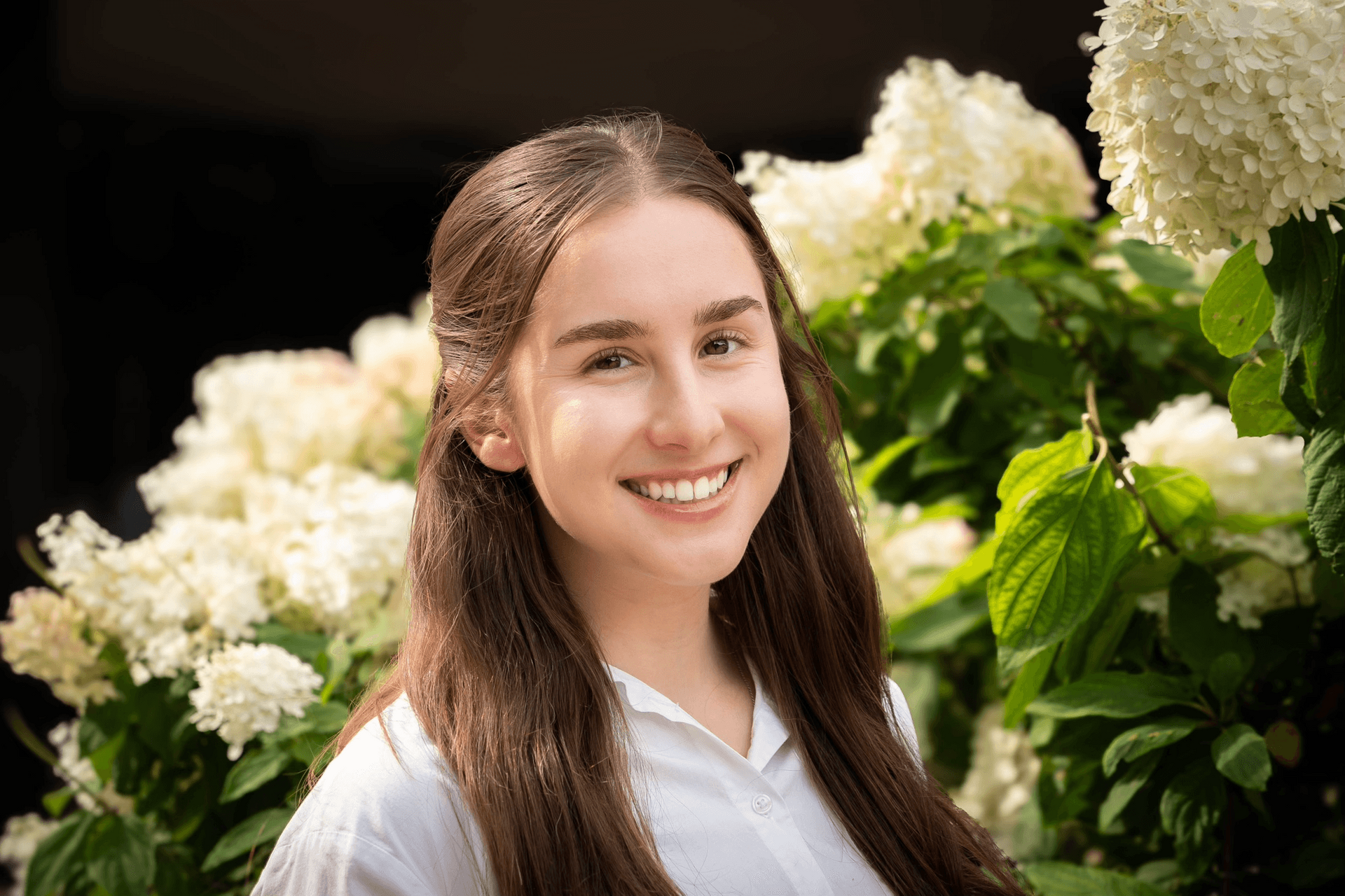 A person with long brown hair, wearing a white shirt, smiles in front of a blooming white hydrangea bush.